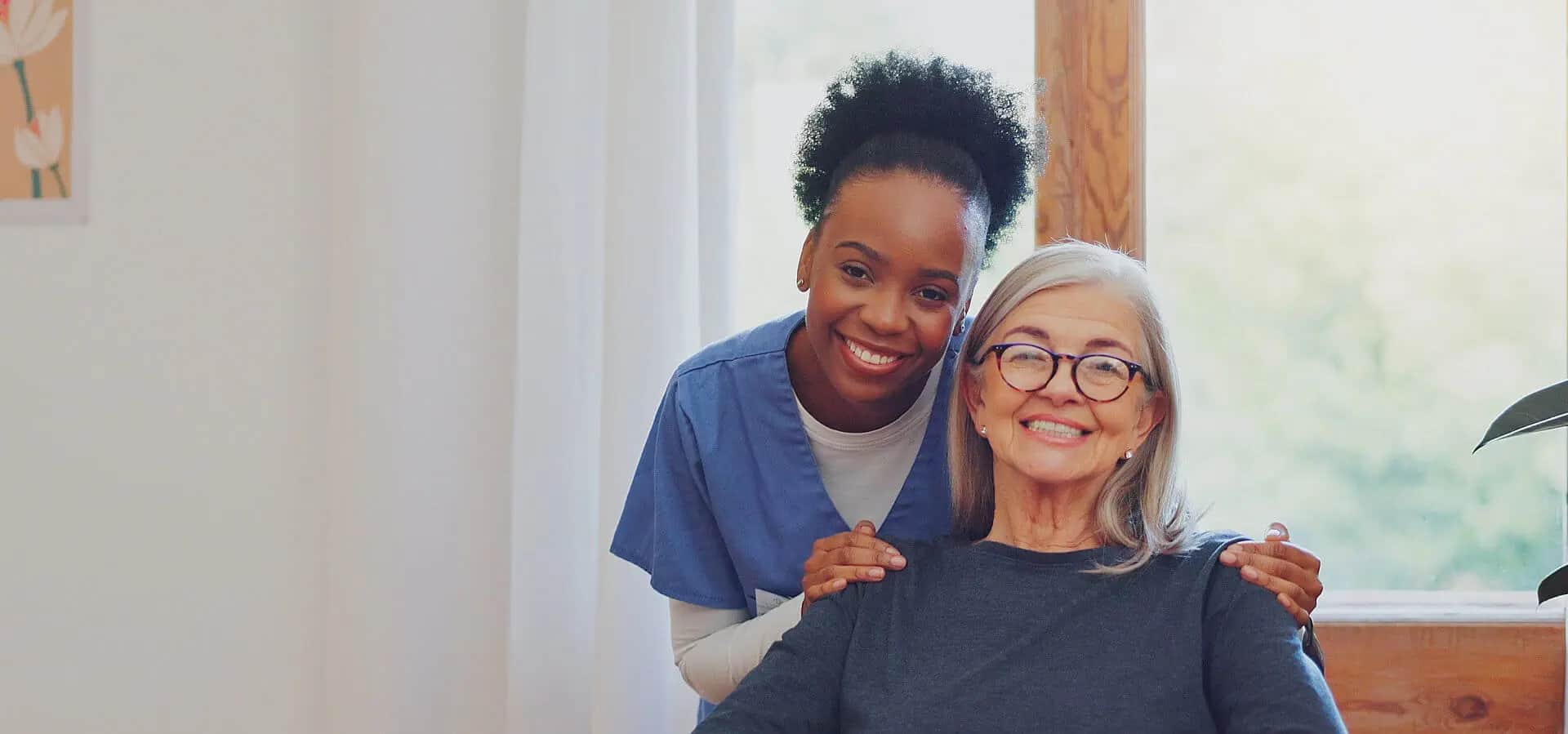Caregiver in scrubs with her hands on a smiling senior woman's shoulders